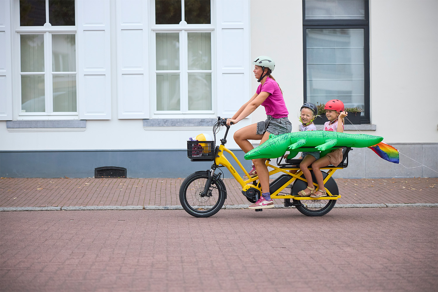 Vrouw fietst met twee kinderen op een gele elektrische longtail fiets door een dorp, met een opblaasbare krokodil achterop.